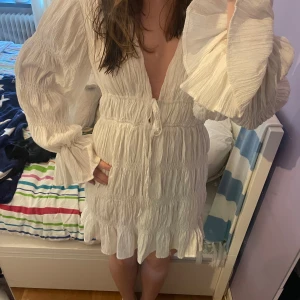 Vit NA-KD klänning  - Säljer en söt vit kortklänning från NA-KD med långa, vida ärmar och volangdetaljer. Klänningen har v-ringning, knyt i midjan och är tillverkad i ett lätt, krinklat material. 