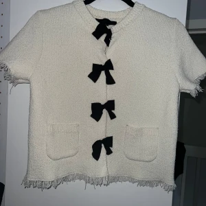 Vit kortärmad kofta med svarta rosetter från Zara - Supersöt vit kofta från Zara med korta ärmar och fyra svarta rosetter framtill. Koftan har två fickor och fransiga kanter för en trendig look. Perfekt att styla till jeans eller kjol!