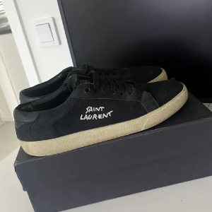 Svarta Saint Laurent sneakers - Svarta sneakers från Saint Laurent med vit broderad logga på sidan. Skorna har svart snörning, rund tå och en beige platt sula. Ovandelen är i canvas och detaljer i skinn, vilket ger en clean och stilren look. Storlek 42 men passar även 43. Hör av er vid frågor. Kan gå ner i pris vid snabb affär!
