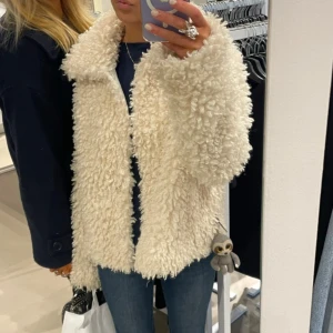 Beige/vit teddyjacka med krage - Supermysig beige/vit teddyjacka med fluffig och lockig yta. Jackan har en bred krage och långärmade ärmar, perfekt för dig som vill ha en trendig och varm look. Passar snyggt till jeans och sneakers för en chill vibe.