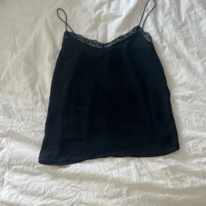 Svart linnetopp med spetskant - Snygg svart linnetopp med tunna axelband och fin spetskant upptill. Perfekt att styla lager på lager eller bära som den är. Toppens material känns lätt och svalt, vilket gör den superbekväm. Enkel men ändå elegant design som passar till många olika looks.