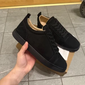 Svarta sneakers från Louboutin mocka - Snygga svarta sneakers från Louboutin i mjuk mocka med klassisk röd sula och svarta snören. Skorna har en stilren design med rund tå och platt sula, samt beige innersida för extra komfort. Perfekta för dig som gillar lyxiga detaljer och streetstyle.