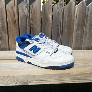 New Balance 550 vita och blå sneakers - Snygga New Balance 550 sneakers i vitt och blått med svarta detaljer. Skorna har klassisk snörning, rund tå och platt sula. Ovandelen är i skinn och textil, med tydlig New Balance-logga på sidan och bak. Perfekta för dig som gillar retrostil och streetwear. ‼️Inga inre skosulor‼️