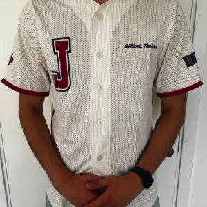 Vit mesh baseballskjorta Florida - Vit baseballinspirerad skjorta i mesh med röda detaljer och stora bokstaven J på bröstet. Kortärmad med knappar framtill och broderad text 'St. River, Florida'. Röd kant vid ärmslut och patch på ärmen. Perfekt för sportig stil.