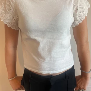 Vit blus med broderade ärmar - Superfin vit blus med korta, broderade ärmar i bomull. Ärmarna har vågig kant och blommigt hålmönster som ger en söt och somrig vibe. Rak passform och rund halsringning. Perfekt att styla med jeans eller kjol för en fräsch look.