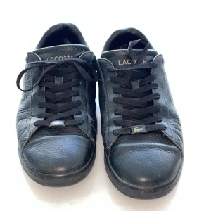 Svarta sneakers från Lacoste i skinn - Klassiska svarta Lacoste sneakers i skinn med låg profil och rund tå. Skorna har snörning och den ikoniska krokodilloggan i silver på sidan. Perfekta för en clean och stilren look. Modell: Carnaby Evo.