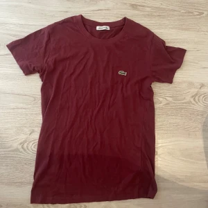 Vinröd Lacoste t-shirt i bomull - Vinröd t-shirt från Lacoste i mjuk bomull med klassisk krokodillogga broderad på bröstet. Enkel och stilren design med rund hals och korta ärmar. Perfekt för dig som gillar en clean och tidlös look.