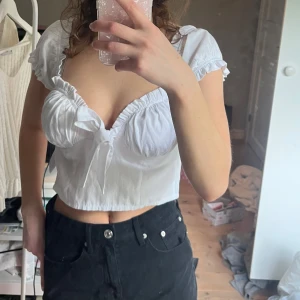 Vit croppad blus med knyt - Supersöt vit croppad blus med korta puffärmar, volangdetaljer och knyt framtill. Djup v-ringning och rynkade detaljer vid bysten ger en romantisk vibe. Perfekt att styla med jeans eller kjol för en trendig look. Toppen har en dragkedja på sidan, då toppen är för liten för mig är dragkedjan öppen. Den sitter alltså lite tightare om den passar din storlek :)