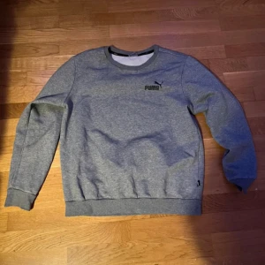 Grå sweatshirt från Puma - Säljer en stilren grå sweatshirt från Puma med svart logga på bröstet. Tröjan har rund halsringning, långa ärmar och ribbade muddar vid ärmslut och nederkant. Perfekt för en chill och sportig look.