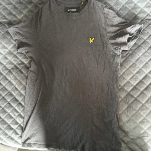 Mörkgrå t-shirt från Lyle & Scott - Snygg mörkgrå t-shirt från Lyle & Scott med klassisk passform och korta ärmar. Bröstet pryds av den ikoniska gula örnloggan. Tillverkad i mjuk bomull som känns skön mot huden. Perfekt för en clean och stilren look.