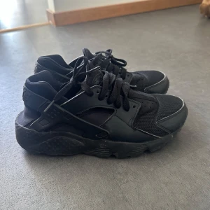 Svarta Nike Huarache sneakers - Säljer ett par helsvarta Nike Huarache sneakers i storlek 38.5. Skorna har en chunky sula, mesh och syntetdetaljer samt snörning. Perfekta för dig som gillar sportig och urban stil. Nike-logga bak på hälen och klassisk design.
