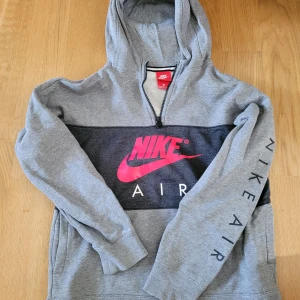Grå Nike Air hoodie med rött tryck - Snygg grå Nike Air hoodie med svart panel och stort rött Nike-tryck på bröstet. Halv dragkedja framtill, huva och fickor. Långärmad med Nike Air-tryck längs ena ärmen. Perfekt för en sportig och avslappnad stil.