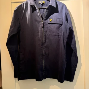 Mörkblå overshirt från Lyle & Scott - Snygg mörkblå overshirt från Lyle & Scott med dragkedja framtill och klassisk krage. Sparsamt använd. Storlek 14/15 år 