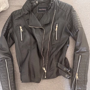 Svart skinnjacka från King of Fashion - Snygg svart skinnjacka med coola detaljer från King of Fashion. Jackan har flera dragkedjor, quiltade partier på axlar och ärmar samt bälte i midjan. Perfekt för dig som vill ha en edgy look och gillar bikerstil.
