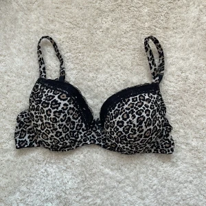 Leopardmönstrad bh från Trofé - Snygg leopardmönstrad bh från Trofé med svarta spetsdetaljer och liten rosett med pärlor framtill. Justerbara axelband och vadderade kupor. Tillverkad i mjuk polyamidblandning för extra komfort.
