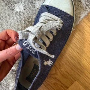 Mörkblå sneakers från Polo Ralph Lauren - Snygga mörkblå sneakers från Polo Ralph Lauren med vit broderad logga på sidan och grova vita snören. Ovandelen har ett diskret blommönster i tyg och tåpartiet är förstärkt med vit gummikappa. Klassisk platt sula med Polo-mönster undertill.