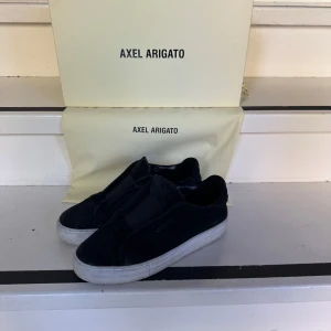 Svarta sneakers från Axel Arigato - Stilrena svarta sneakers från Axel Arigato med vit platt sula och minimalistisk design. Skorna är i mocka och har elastiskt band över vristen istället för klassisk snörning. Perfekta för dig som gillar en clean och modern look. Passar storlek 40