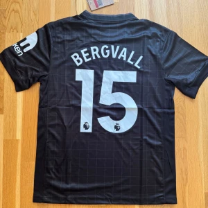 Tottenham Bergvall #15 svart matchtröja - Snygg svart Tottenham Hotspur matchtröja från Nike med Bergvall och nummer 15 på ryggen. Tröjan har diskret rutmönster, korta ärmar och klassisk rund hals. Materialet är lätt och andas, perfekt för fotboll eller häng. AIA-logga på bröstet och Premier League-detaljer.