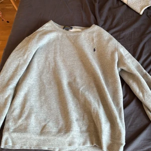 Grå sweatshirt från Polo Ralph Lauren - Klassisk grå sweatshirt från Polo Ralph Lauren med broderad marinblå logga på bröstet. Tröjan har rund halsringning och långa ärmar, perfekt för en chill och stilren look. Tillverkad i mjuk bomull för maximal komfort.