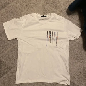 Vit AMIRI t-shirt med färgstänk - Vit t-shirt från AMIRI med korta ärmar och coolt tryck både fram och bak. Stora AMIRI-bokstäver med färgstänk och drip-effekt i blått, orange och svart. Skön passform och klassisk rund hals. Perfekt för dig som gillar streetstyle och vill sticka ut.