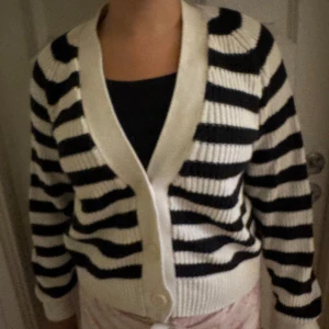 Randig kofta från Lindex i svart och vitt - Säljer en ribbstickad kofta från Lindex i svart och vitt med breda ränder. Koftan har v-ringning, stora knappar framtill och långa ärmar. Perfekt för dig som gillar en chill och klassisk look med lite edge.