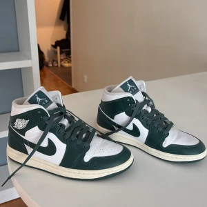 Nike Air Jordan 1 Mid vit/grön - Nike Air Jordan 1 Mid sneakers i vitt och mörkgrönt skinn. Klassisk siluett med snörning, rund tå och ikoniska Jordan Wings-loggan på sidan. Snygg kontrast mellan de gröna och vita panelerna, perfekt för dig som gillar streetstyle och sneakers med retrovibe.
