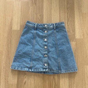 Jeanskjol med knappar från H&M - Snygg blå jeanskjol från Divided med A-linjeform och knappar framtill. Kjolen har bälteshällor och klassisk jeanssöm. Perfekt för dig som gillar en enkel men trendig stil.