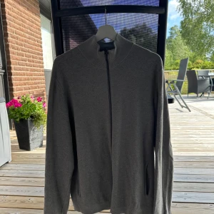 Mörkgrå kofta med dragkedja - En stilren mörkgrå kofta med hel dragkedja framtill och hög krage. Tröjan har långa ärmar och en enkel, clean design som passar perfekt till lager-på-lager. Materialet känns mjukt och bekvämt, perfekt för kyligare dagar.