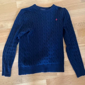 Mörkblå kabelstickad tröja Morris - Säljer en mörkblå kabelstickad tröja från Morris med rund hals och röd logga broderad på bröstet. Tröjan har långa ärmar och ribbade muddar. 