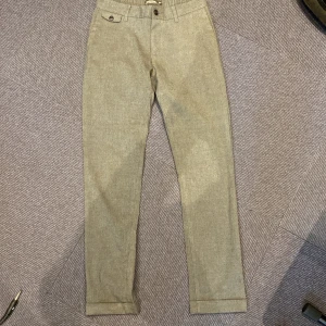 Grå kostymbyxor Dressmann slim fit - Snygga grå kostymbyxor från Dressmann i slim fit-modell. Byxorna har klassisk design med fickor fram och bak, knappdetaljer och uppvikta benslut. Perfekta för en clean och stilren look.