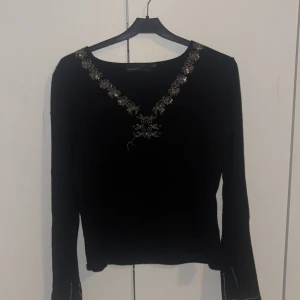 Svart tröja med broderi från KappAhl - Svart långärmad tröja från KappAhl med snyggt broderi i silver och guld längs v-ringningen och vid ärmsluten. Tröjan har en klassisk passform och är perfekt för dig som gillar detaljer. Superfin att styla med jeans eller kjol. Använd 1 gång 