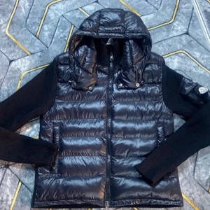 Svart Moncler pufferjacka med huva - Snygg svart pufferjacka från Moncler med glansig framsida och stickade ärmar. Jackan har huva, dragkedja och Moncler-logga på ärmen. Perfekt för dig som vill ha en sportig och trendig look med både värme och stil.