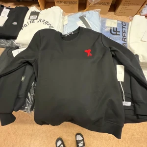 Svart sweatshirt från Ami Paris - Svart sweatshirt från Ami Paris med röd broderad logga i form av ett hjärta och bokstaven A på bröstet. Tröjan har rund halsringning och är tillverkad i 100% bomull. Perfekt för en clean och stilren look.