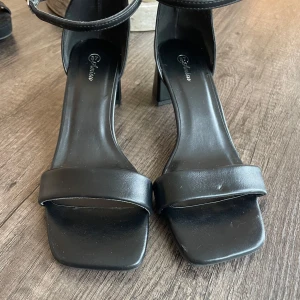 Svarta sandaler med klack från Anisis - Stilrena svarta sandaler från Anisis med fyrkantig tå, bred rem över foten och justerbart ankelband. Skorna har en stabil blockklack och är tillverkade i syntetmaterial med en matt finish. Perfekta för dig som gillar minimalistisk och trendig look. Stl 37. Jag har vanligtvis 38 men fick ta 37 så dem är lite stora i storlek. Endast använda 1 gång 