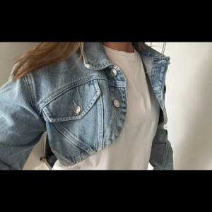 Croppad jeansjacka från Zara - Snygg croppad jeansjacka från Zara i ljusblå denim. Klassisk krage, silverfärgade knappar och två bröstfickor med lock. Jackan har lång ärm och en loose passform som ger en chill vibe till outfiten.
