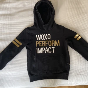 Svart hoodie från WOXO med guldtryck - Svart hoodie från WOXO med texten 'WOXO PERFORM IMPACT' i vitt och guld på bröstet. Guldiga ränder på båda ärmarna, huva och två dragkedjefickor framtill. Perfekt för dig som gillar sportig och cool stil.