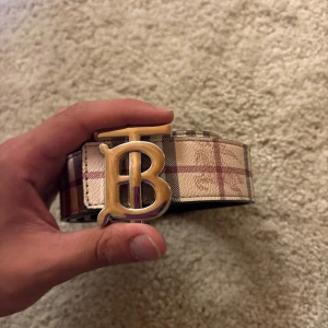 Burberry vändbart bälte med TB-spänne - Säljer ett vändbart Burberry-bälte med ikoniskt TB-spänne i metall. Ena sidan har klassiskt rutigt mönster i beige, brun och röd, andra sidan är svart med präglade detaljer. Bältet är brett och har lyxig känsla, perfekt för att lyfta din outfit.👍