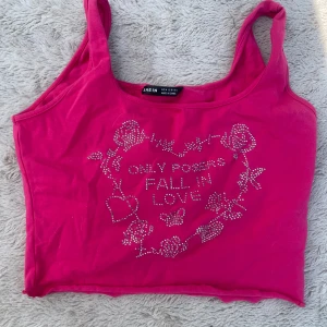 Rosa croppad linne från SHEIN - Snyggt rosa croppat linne från SHEIN med glittriga stenar framtill som bildar texten 'Only posers fall in love' och dekorationer med rosor, hjärta och fjäril. Mjuk bomullskänsla och breda axelband, perfekt för en färgstark look.