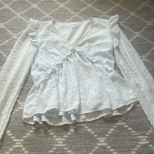 Ljusblå blus med volang från Gina Tricot - Supersöt ljusblå blus från Gina Tricot med v-ringning, volangdetaljer och långärmad design. Tunt, lätt tyg med små prickar i strukturen som ger en extra touch. Perfekt för dig som gillar romantisk och trendig stil.