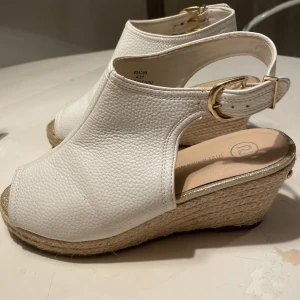 Vita skor med kilklack och spänne - Snygga vita sandaler från River Island med grov textur i skinnimitation, öppen tå och justerbart guldfärgat spänne. Kilklack i flätad jute ger en somrig vibe och extra höjd. Perfekta för att lyfta din outfit med en fräsch touch.