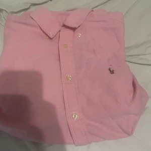 Ljusblå skjorta från Polo Ralph Lauren - Klassisk ljusblå skjorta från Polo Ralph Lauren med button-down krage och broderad logga på bröstet. Skjortan har vita knappar och är tillverkad i mjuk bomull. Perfekt för en clean och stilren look.