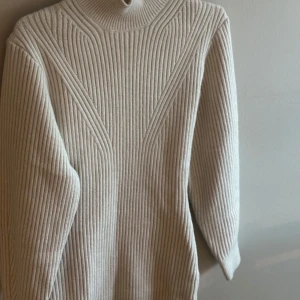 Beige ribbad klänning från H&M - Säljer en beige ribbad klänning från H&M i storlek XS. Klänningen  har lång ärm, hög krage och är stickad i ett mjukt material. Snyggt mönster med vertikala och diagonala ränder som ger en stilren look.