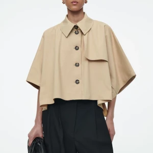 Beige croppad trenchcoat från COS - Säljer en beige croppad trenchcoat från COS med stora knappar framtill och bred krage. Jackan har vida, korta ärmar och en asymmetrisk detalj på ena axeln. Perfekt för dig som gillar minimalistisk och trendig stil. Slutsåld och orginalpris 1300kr