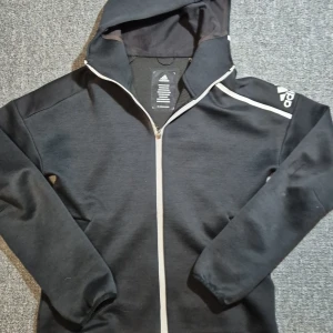 Svart Adidas hoodie med dragkedja - Svart hoodie från Adidas med vit dragkedja och vita detaljer på axeln. Jackan har huva, två fickor och Adidas-logga på ärmen. Perfekt för dig som gillar sportig stil och vill ha något bekvämt och snyggt till vardags.