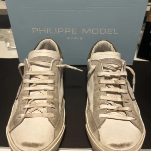 Philippe Model  - Snygga vita sneakers från Philippe Model med detaljer i beige mocka och klassisk logotyp på sidan. Skorna har snörning, platt sula i gummi och rund tå. Perfekta för dig som gillar stilrena och exklusiva sneakers med en touch av Paris. Och man får såklart med box.🤩