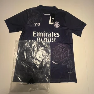 Real Madrid x Y-3 Adidas fotbollströja - Snygg marinblå Real Madrid fotbollströja från Adidas och Y-3 med vita tryck, klubbmärke och Emirates Fly Better-logga. Unik rosdetalj på framsidan och baksidan. Tillverkad i lätt funktionsmaterial (AEROREADY) och har korta ärmar.