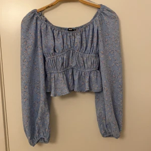 Blommig blå blus från BikBok - Supersöt blommig blus i ljusblått från BikBok. Modellen är croppad med volangkant nertill, smockad midja och långa puffärmar med resår. Blusen har offshoulder-skärning och små blommor i rosa och vitt över hela tyget. Perfekt för vår och sommar!
