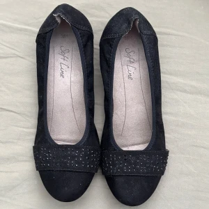 Svarta ballerinaskor från Soft Line - Stilrena svarta ballerinaskor från Soft Line med rund tå och dekorativa svarta nitar framtill. Skorna är gjorda i mocka-liknande material och har en platt sula som gör dem bekväma för vardagsbruk. Perfekta att matcha med både jeans och klänning.