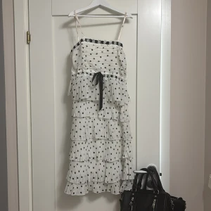 Vit prickig volangklänning med rosett - Supersöt vit kortklänning med svarta prickar och flera volanger. Klänningen har smala axelband, svart rosett i midjan och dekorativ spets upptill. Perfekt för dig som gillar lekfull och trendig stil. Klänningen har tyvärr små fläckar på sig här och där, men det är inget som syns om dessa går förmodligen bort i tvätten☺️