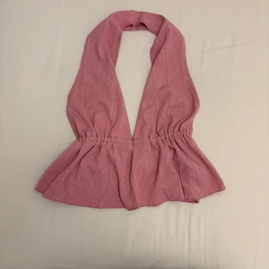Rosa halterneck topp med djup v-ringning - Supersöt rosa omlott topp med djup v-ringning och markerad midja. Toppen är ärmlös och har en lätt utsvängd nederdel som ger en snygg siluett. Perfekt för dig som vill sticka ut med en feminin och trendig look.